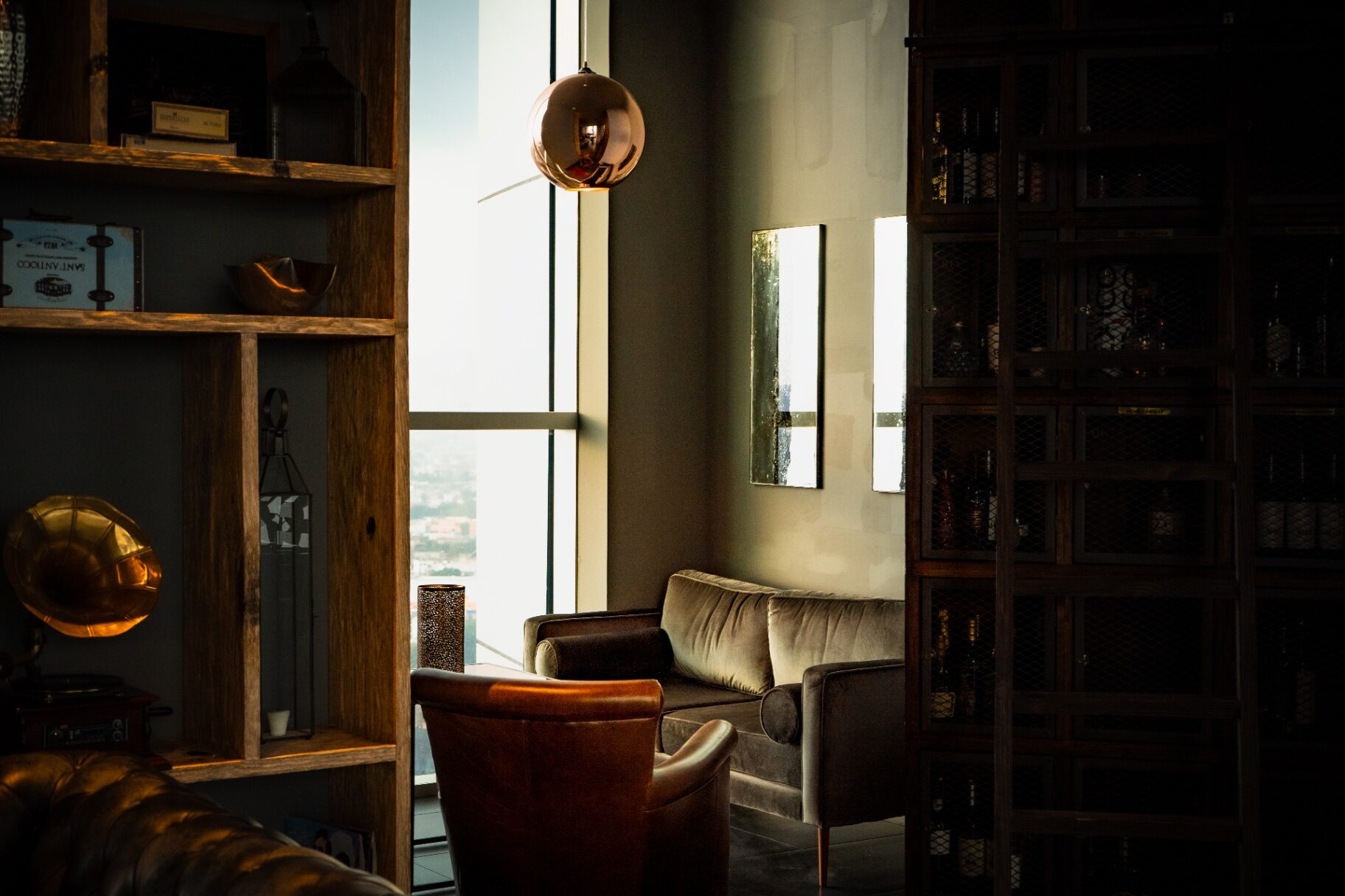 Salón privado en penumbra con chesterfield de piel, sofá de terciopelo, lámpara esférica de cobre y biblioteca de maderas oscuras al atardecer.