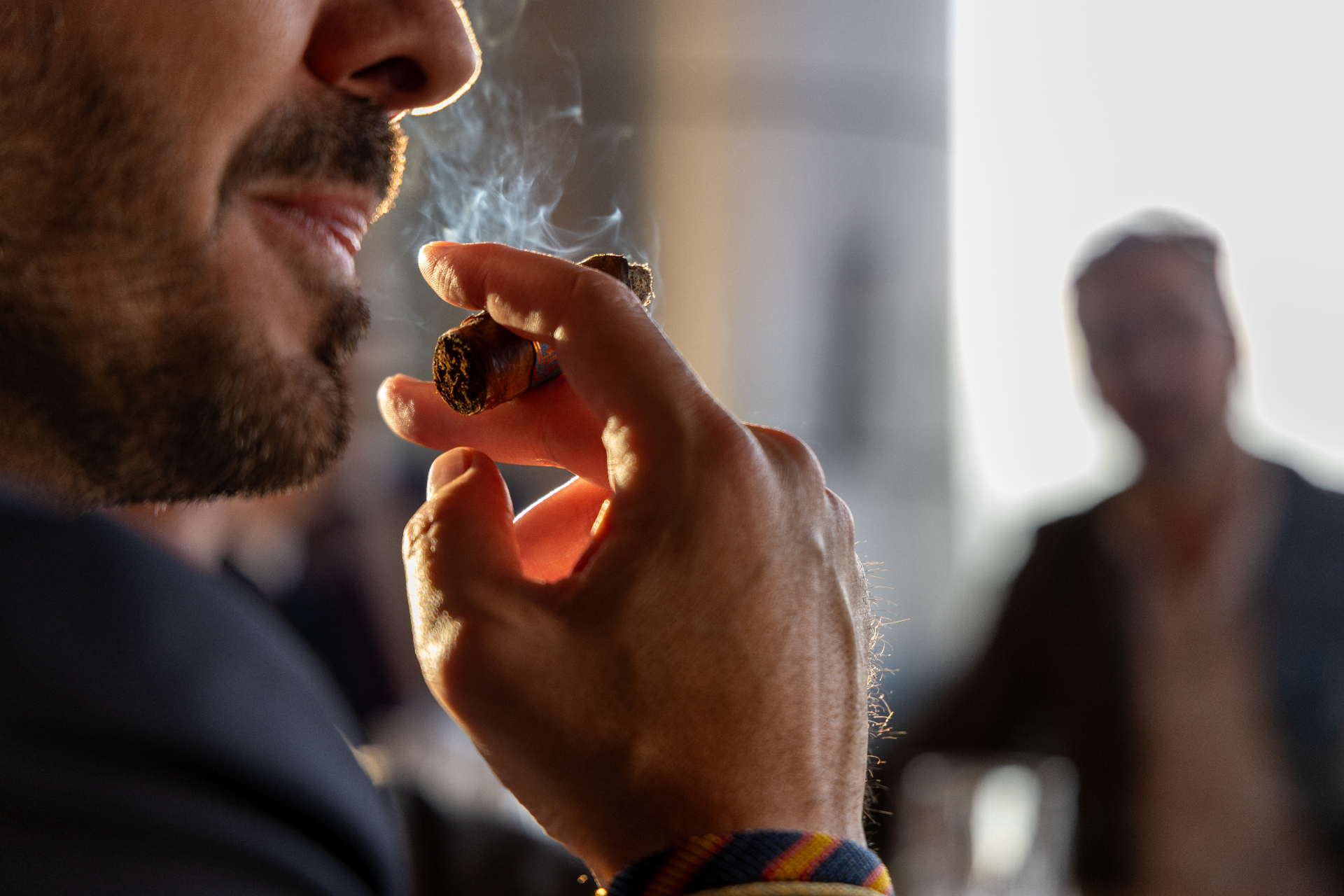 Primer plano de una mano sosteniendo un puro con humo suave y luz cálida de fondo, evocando la atmósfera de un club privado.