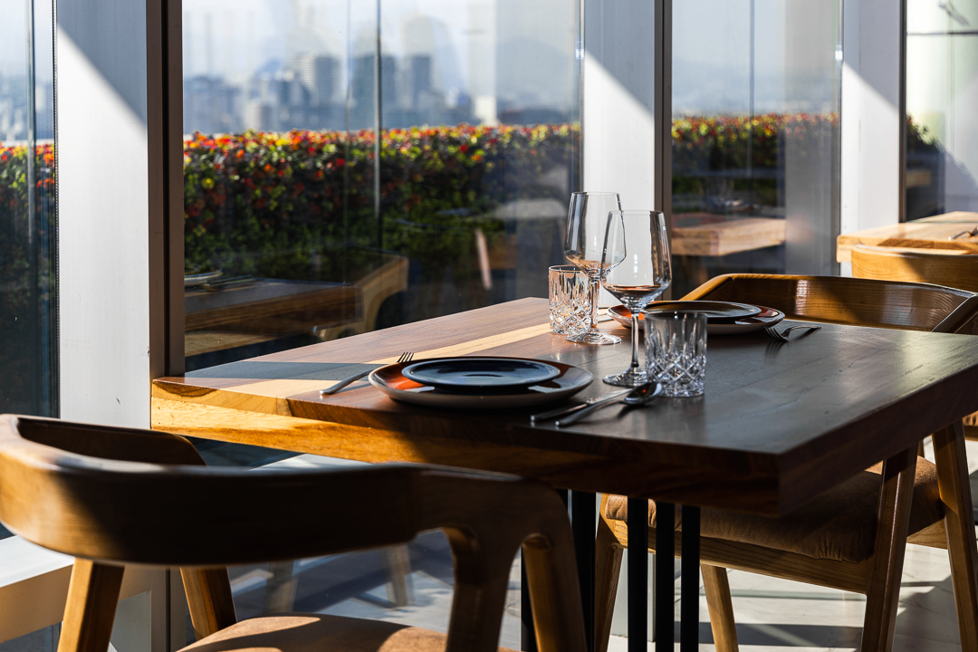 Mesa de comedor con cristalería bajo luz natural, frente a ventanales de piso a techo con la Ciudad de México al fondo.