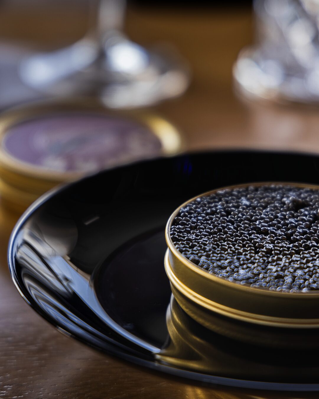 Lata abierta de caviar servida sobre un plato negro lacado con cristalería desenfocada al fondo.