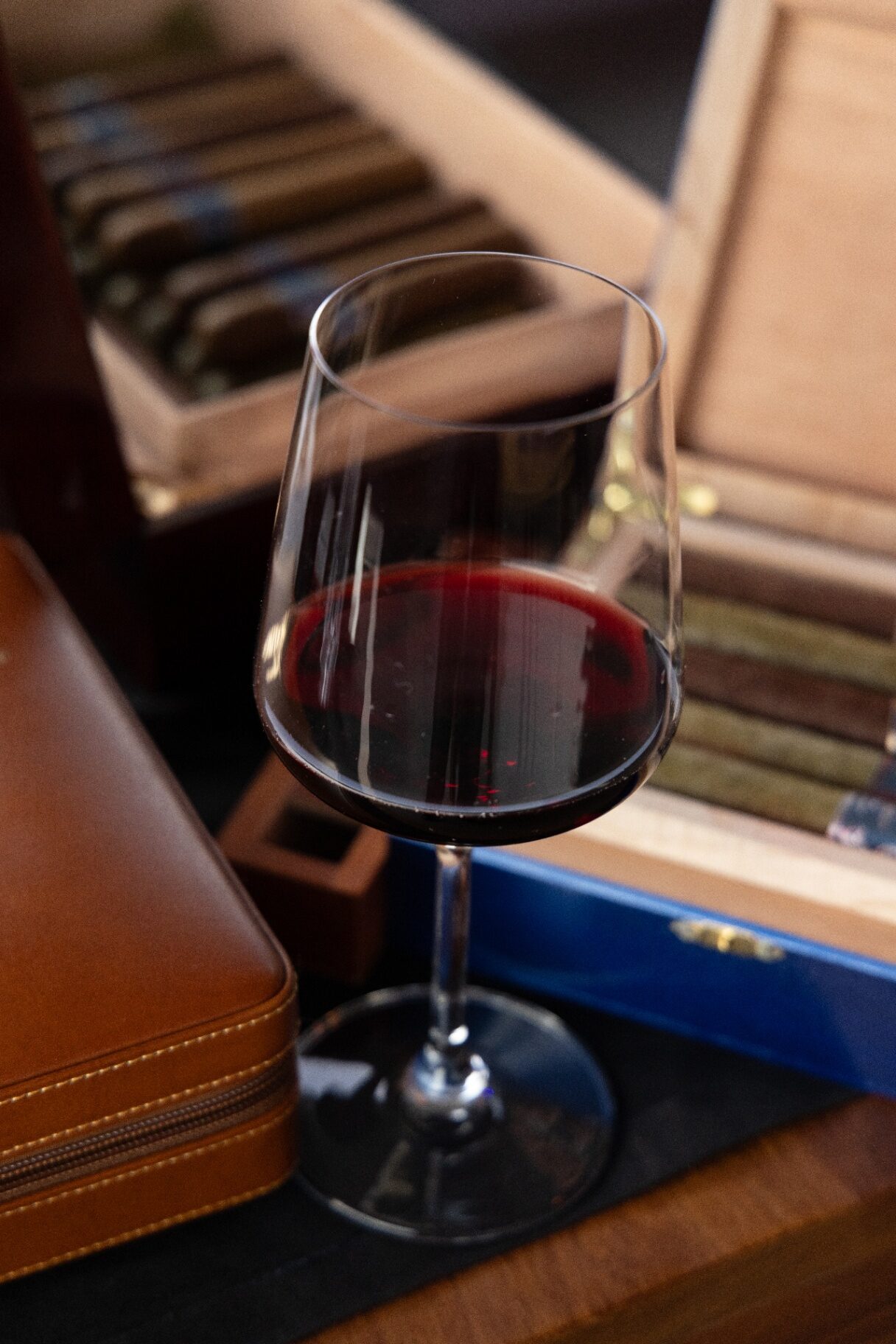Copa de vino tinto junto a un humidor abierto y un estuche de piel, evocando el arte de vivir y las colecciones privadas.
